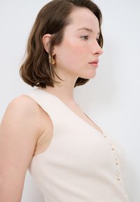 Gestreifter beiger Tanktop mit tiefem V-Ausschnitt und goldenen Knöpfen auf der Vorderseite. Der Stoff wirkt weich und leicht strukturiert.