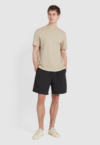 T-shirt beige a maniche corte con scollo tondo e piccolo emblema sul petto, abbinato a pantaloni corti neri e sneakers di colore chiaro.