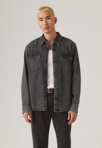 Camisa de denim negra con cierre de botones, dos bolsillos en el pecho y mangas largas. Llevada sobre una camiseta sin mangas blanca, combinada con pantalones oscuros.