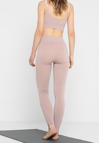 Kobieta stojąca boso na macie do jogi, ubrana w mauve wyższe legginsy i dopasowany stanik sportowy, zwrócona plecami na jednolitym tle.