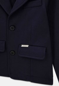 Blazer bleu marine foncé avec deux boutons noirs et poches à rabat, dont une poche ornée d'une plaque avec le logo argenté "GUESS".