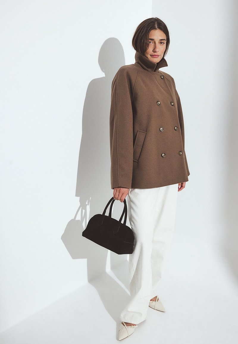 Brūns dubultās krūšu mētelis ar augstu apkakli, teksturētu audumu un sudraba pogām; kopā ar baltām plašām biksēm un mazu melnu rokassomu.