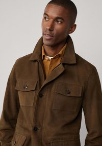 Suede-braune Jacke mit vier Vorderaschen, schwarzen Knöpfen und einem klassischen Kragen. Getragen über einem senfgelben Hemd mit glatter Textur.