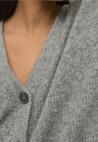 Cárdigan gris de punto con un diseño de cuello en V, que presenta un botón negro prominente. Textura suave y patrón sutil visible en la tela.