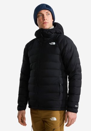Man som bär en svart vadderad North Face-jacka, blå stickad mössa och bruna byxor, tittar något åt sidan mot en vit bakgrund.