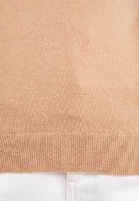 Ourlet de pull beige tricoté sur pantalon blanc, montrant les points et la texture du tissu en gros plan.