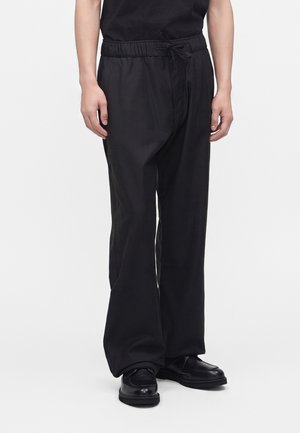 Homme portant un pantalon ample noir à cordon avec des chaussures en cuir noir sur fond blanc.