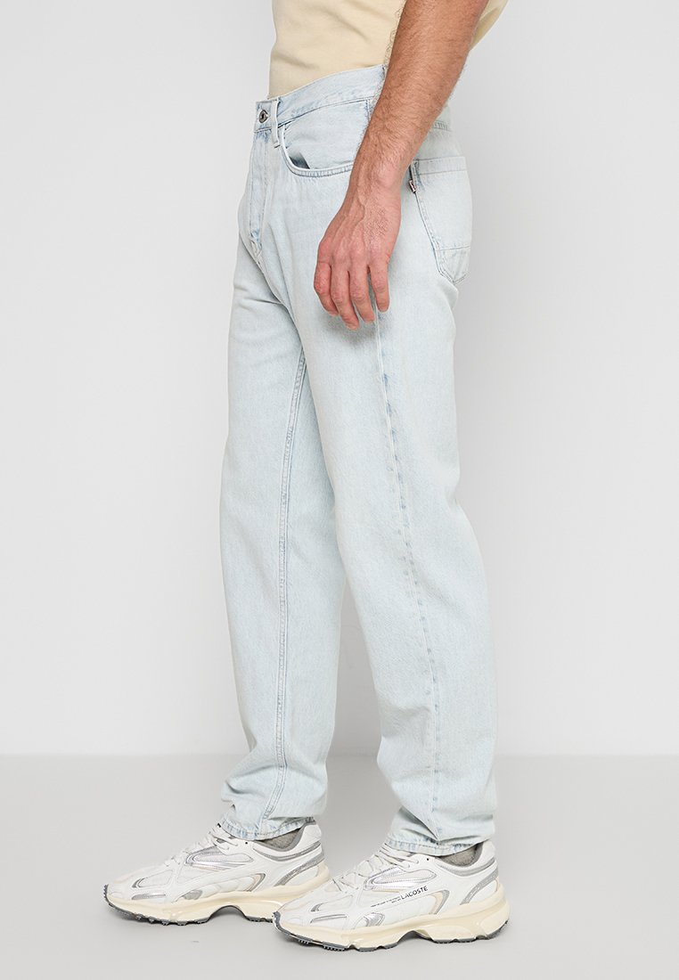 Hellblaue Jeans im Relaxed Fit mit fünf Taschen und einem klassischen Knopfverschluss. Getragen mit weißen Sportschuhen.