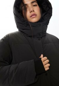 Crna puffa jakna s kapuljačom, s prošivenim dizajnom, dugmadi na kopčanje i podesivim vezicama. Ima rebraste manžetne za dodatnu toplinu.