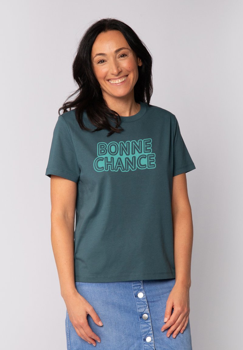 T{e-}al Baumwoll-T-Shirt mit kurzen Ärmeln, das den Text "BONNE CHANCE" in fetten, umrandeten Buchstaben zeigt. Kombiniert mit einem Jeansrock.