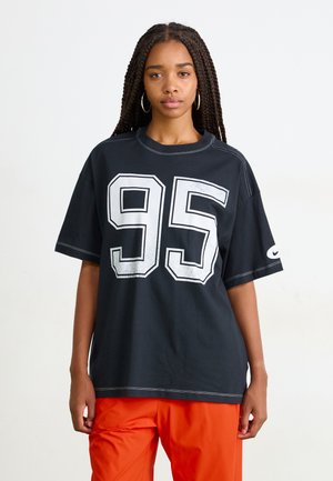 T-shirt noir avec un grand imprimé blanc "95", manches courtes, col rond et détails de couture contrastants. Associé avec un pantalon orange.