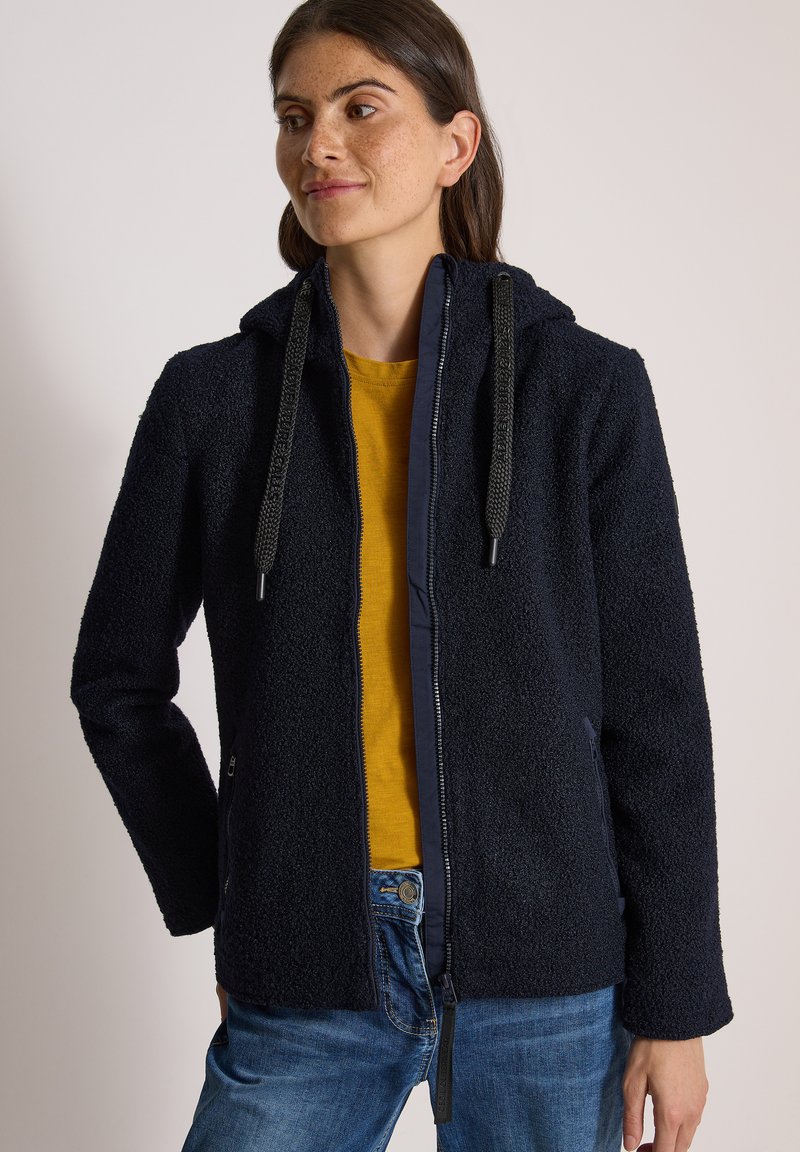 Femme portant une veste polaire zippée bleu marine avec capuche et larges cordons noirs sur une chemise jaune moutarde et un jean bleu.