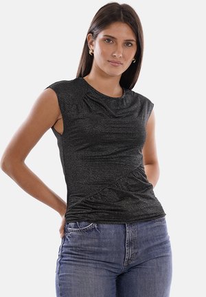 Donna con capelli lunghi e scuri che indossa un top nero senza maniche con texture e jeans blu, in piedi con una mano dietro la schiena.