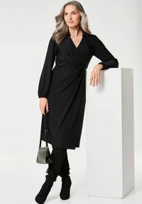 Schwarzes Langarmkleid mit Wickeldesign, ausgestattet mit einem tiefen V-Ausschnitt und einer Raffung an der Taille. Kombiniert mit hohen schwarzen Wildlederstiefeln und einer kleinen Tasche.