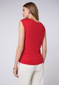 Haut sans manches rouge avec un décolleté arrondi et une texture lisse, associé à un pantalon de couleur claire. Design simple sans détails supplémentaires.