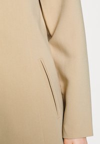 Giacca beige con una texture liscia, maniche lunghe e una tasca laterale con dettaglio di cucitura. Il tessuto appare morbido e ben adattato.