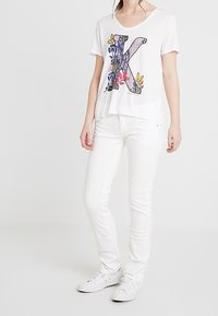 Camiseta gráfica blanca con un diseño floral en 'K' azul y multicolor, combinada con jeans ajustados blancos y zapatillas blancas.