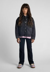 Chaqueta de mezclilla en un lavado oscuro con cierre de botones y bolsillos frontales, superpuesta sobre una blusa rosa claro y combinada con pantalones negros y zapatillas de colores.