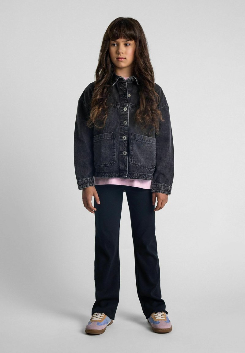 Chaqueta de mezclilla en un lavado oscuro con cierre de botones y bolsillos frontales, superpuesta sobre una blusa rosa claro y combinada con pantalones negros y zapatillas de colores.