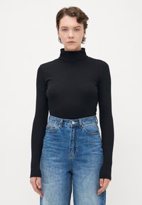 Woman with short wavy hair wears black long-sleeve turtleneck and high-waisted blue jeans, standing against plain white background.