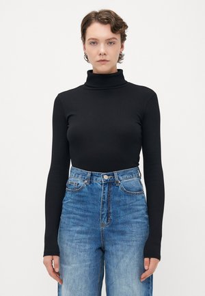 Kvinne med kort bølget hår har på seg svart langermet genser med høy krage og høy midje blå jeans, står mot en enkel hvit bakgrunn.