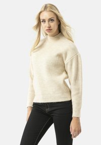 Beige ribbeltrui met hoge hals, ontspannen pasvorm en verlaagde schouders. Gepaard met donkere jeans met contrasterende stiksels.