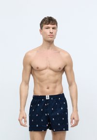 Mørkeblå boxershorts i åndbart bomuld med et gentaget mønster af små farvede motiver. Har en elastisk talje og knaplukning.