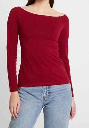 Frau trägt ein langärmeliges, schulterfreies, eng anliegendes rotes Oberteil kombiniert mit hellblauen High-Waist-Jeans vor einfachem Hintergrund.