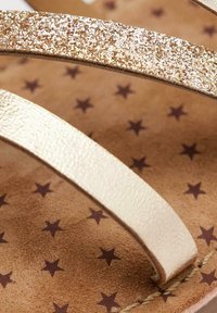 Sandali infradito dorati con glitter, con una suola piatta e testurizzata di colore marrone che presenta piccole stelle scure. Cinghie sottili, una lucida e una opaca.