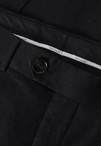 Pantalones de tela negra con frente plano, cierre de botón y una textura sutil. La etiqueta interior dice "Tantuum". Enfoque en líneas limpias y diseño clásico.