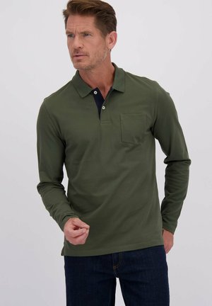 Homme portant un polo à manches longues olive et un jean foncé, se tenant contre un fond clair uni, regardant sur le côté.