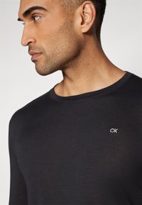 Svart langermet skjorte laget av lett stoff med rund halslinning, med et lite hvitt brodert "CK"-logo på brystet.