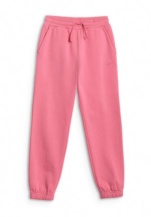 Fille aux longs cheveux portant un bonnet rose, un pantalon de survêtement rose, un crop top blanc et des baskets blanches, debout les mains derrière la tête.