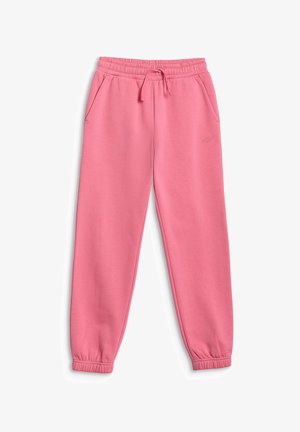Fille aux longs cheveux portant un bonnet rose, un pantalon de survêtement rose, un crop top blanc et des baskets blanches, debout les mains derrière la tête.