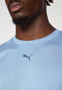 Lyseblå idretts-T-skjorte med rund hals og en mørkeblå Puma-logo sentrert på brystet. Glatt, lett stoffkvalitet.