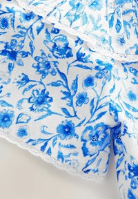 Weißer Stoff mit einem blauen Blumenmuster, das verschiedene Blumen zeigt. Mit zarter Spitzenkante verziert. Sanfte Textur, leichtes und luftiges Design.