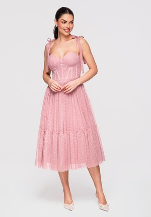 Femme debout et souriante, portant une robe bustier en dentelle rose avec des bretelles à nouer et une jupe en tulle à pois à volants, associée à des talons blancs.