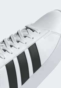 Zapatilla blanca con diseño de tres franjas negras, cordones blancos y parte superior de cuero texturizado.