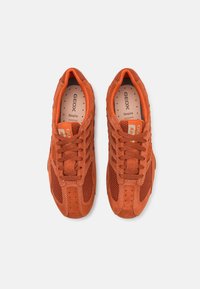 Orange suede sneakers with mesh detailing, featuring round laces, perforations for ventilation, and branding on the tongue and heel.