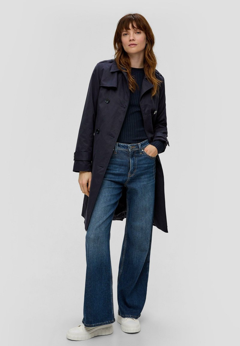 Dunkelblauer Trenchcoat über einem gerippten dunkelblauen Oberteil, kombiniert mit hoch taillierten, weit geschnittenen blauen Jeans und weißen Sneakers. Verfügt über doppelte Knöpfe.