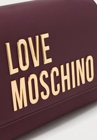 Bordeaux faux-lederen portemonnee met gedurfde, metalen gouden opdruk "LOVE MOSCHINO" aan de voorkant. Gladde textuur, rechthoekige vorm.