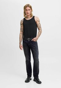 Joven con cabello largo y tatuajes en ambos brazos, vistiendo una camiseta sin mangas negra, jeans negros con cinturón y zapatos negros, de pie frente a un fondo blanco.