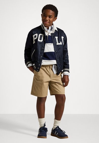 Ναυτική σατινένια μπomber τζάκετ με γράμματα "POLO", ριγέ πουκάμισο, χακί βερμούδες και μπλε αθλητικά παπούτσια με λευκές λεπτομέρειες και γκρίζες κάλτσες.