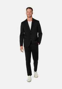Schwarzer Anzug mit einreihigem Sakko und passender Hose. Kombination mit weißem T-Shirt und lässigen weißen Turnschuhen. Glatte Textur.