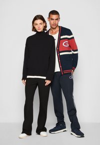 Mujer con un suéter de cuello alto negro; el hombre lleva puesta una chaqueta de punto a rayas en azul marino y rojo con el logo 'G', una camiseta blanca y pantalones de chándal en azul marino. Ambos usan zapatillas deportivas.