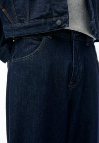 Mørk denimjakke med gyldne accenter, der viser et nærbillede af stoffets tekstur og detaljer på lommerne og sømmene.