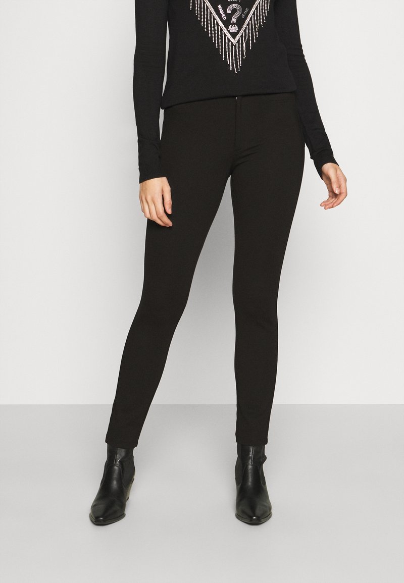 Femme portant un pantalon noir slim, des bottines noires et un top noir à manches longues avec un motif décoratif argenté sur fond blanc.