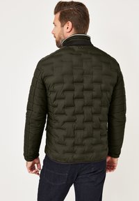 Man with brown hair wearing a dark green quilted jacket and dark blue jeans, facing away against a plain white background.