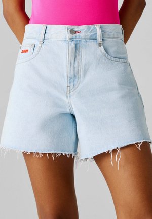 Lyseblå denimshorts med frynser i kanten, båret med en knaldrød top, vist forfra fra taljen til midt på låret.