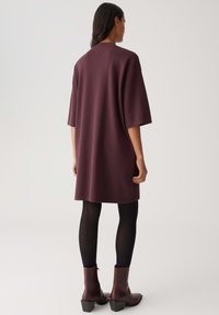 Vrouw met haar rug naar de camera gekeerd, gekleed in een losvallende, knie-lange paarse jurk met driekwart mouwen, zwarte panty en donkerbruine enkellaarsjes met hakken.